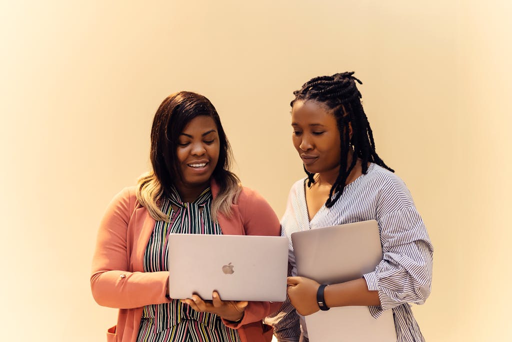 Two women of color stand shoulder to shoulder, both looking down at their laptops. Find more afrocentric images on www.ninthgrid.com